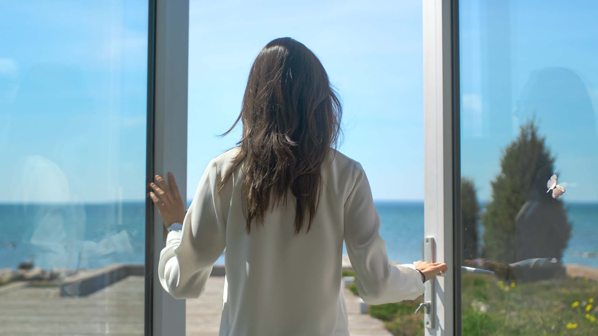 Back of brunette woman in white shirt opening glass doors and walking towards the sea.